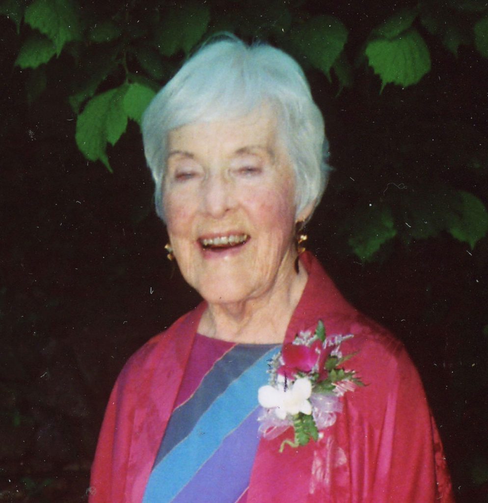 A woman with short, grey hair and dangly earrings smiling brightly wearing a blue and pink patterned blouse, a pink cardigan and a white boutonnière with a dark background with green leaves.