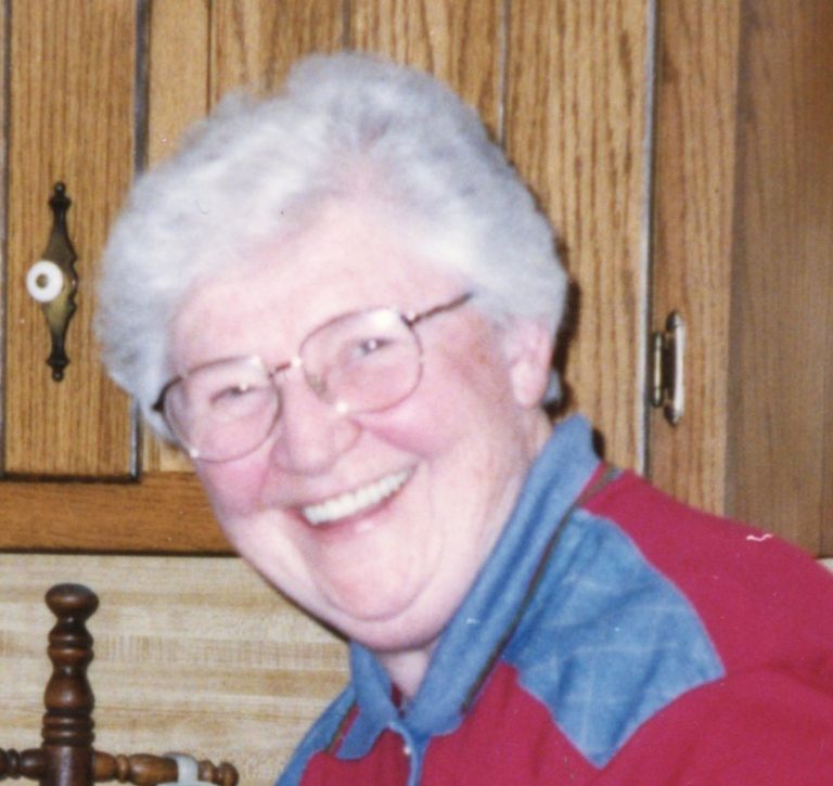 A woman with short, white and grey hair and wire glasses smiling brightly for a headshot picture wearing a red and blue quarter zip in a kitchen with beige wooden cabinets in the background.