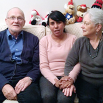 A young woman sitting in between an older man and woman on a couch with Christmas-themed stuffed animals sitting above them on the edge of the couch.