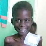 A young girl orphan who is malnourished with medical supplies and tubes taped to her chest. She is standing tall and smiling brightly.