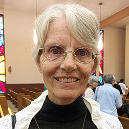 A woman with short white hair and wire-framed glasses smiling brightly for a headshot picture inside a church.
