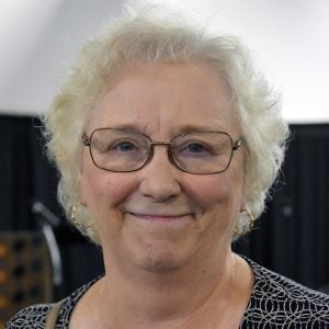 A woman with short white hair and wire-framed glasses smiling brightly for a headshot picture indoors.