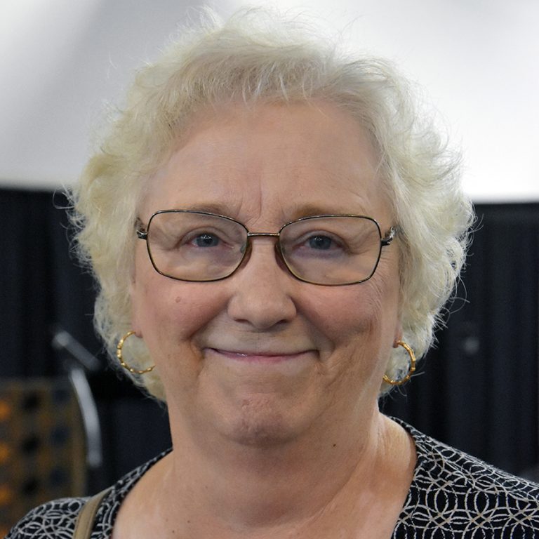 A woman with short white hair and wire-framed glasses smiling brightly for a headshot picture indoors.