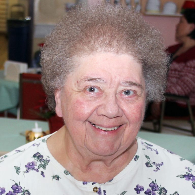 A woman smiling brightly indoors, wearing a white blouse patterned with little purple flowers.
