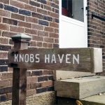 A wooden sign with white lettering that says: knobs haven out front of a red brick building with a white door.