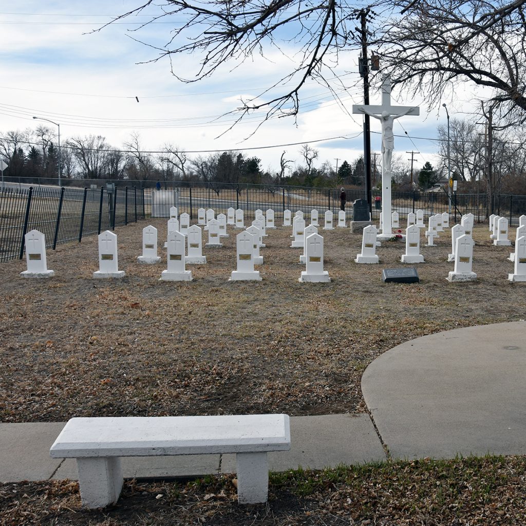 A religious cemetery with white tombstones and a white cross and bench on a sunny day.