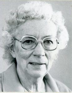 A black and white portrait picture of a woman smiling with short white hair and round glasses with a light colored collared shirt.