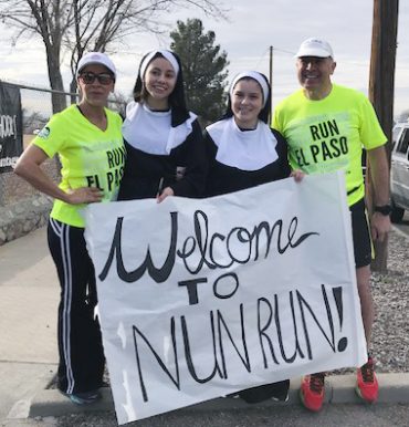 Two nuns and a man and woman wearing neon yellow shirt, white baseball caps, and sunglasses smiling together for a group picture outdoors while holding a large white sign with black lettering that says: Welcome to Nun Run!