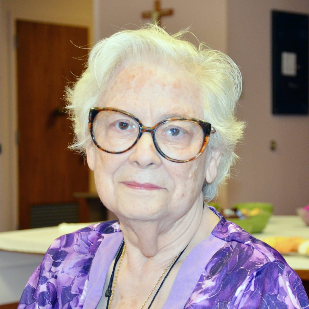 A woman smiling gently with short, white hair, big glasses, and a purple patterned blouse indoors.