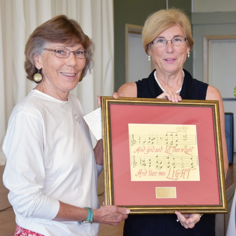 Two women smiling together for a picture in dress clothes while displaying a framed piece of sheet music indoors.