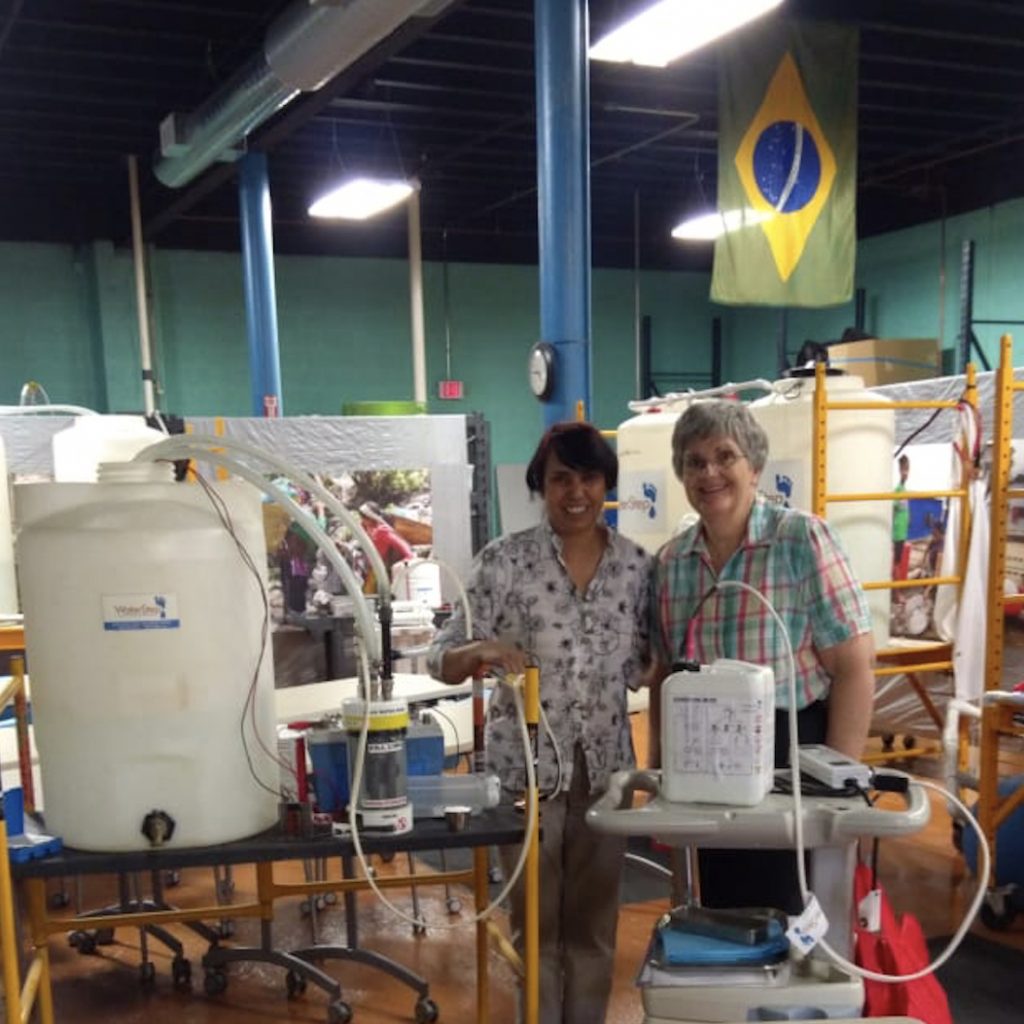 Two women smiling together inside a swimming pool pump room with various equipment, pipes, and tubes.