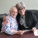 Two women with short grey hair and glasses embracing each other and smiling for a picture together while sitting on a couch and looking at papers together at a table.