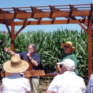 A small crowd of people watching two people speak on a small outdoor stage in front of a tall grass field.