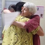 Two women hugging each other indoors. The woman facing the camera has papers in her hands wrapped around her friend.