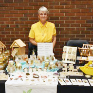 A woman with short grey hair wearing a collared yellow blouse smiling brightly in front of a display table with numerous goods in front of a red brick building indoors.