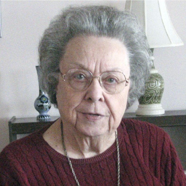 A woman with short, grey hair and glasses smiling for a picture wearing a burgundy sweater, and a necklace indoors.