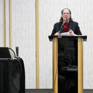 A woman in dress clothes speaking at a social event while a man sitting at a desk to her right watches.