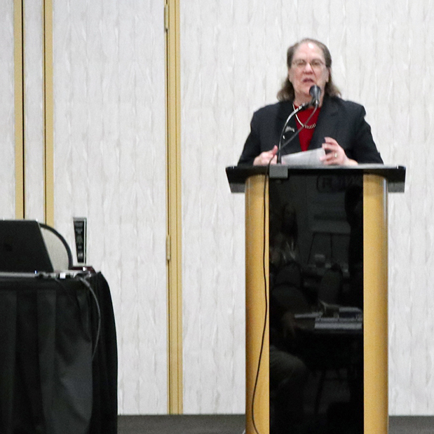 A woman in dress clothes speaking at a social event while a man sitting at a desk to her right watches.