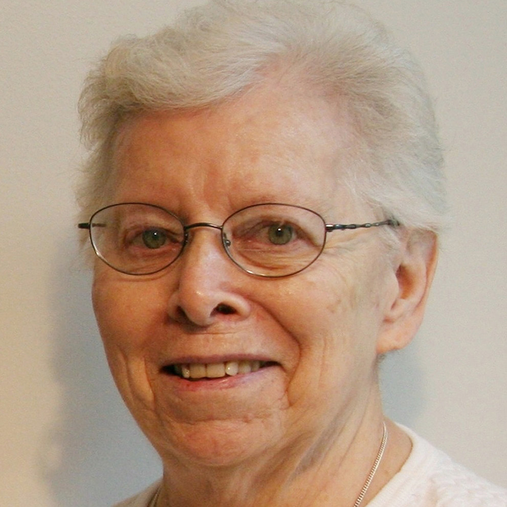 A woman smiling for a headshot picture with short, white hair, and round wire glasses, wearing a white shirt, in front of a plain white background.