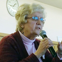 A woman with short grey hair and wire-framed glasses speaking passionately into a microphone at an indoor event.