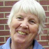 A woman, Maureen Smith, with short white hair, wearing a blue collared shirt, smiling brightly in front of a red brick wall.