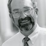 A black and white portrait picture of a man smiling brightly with a beard, and short hair and glasses wearing a white collared shirt and a patterned dark tie.