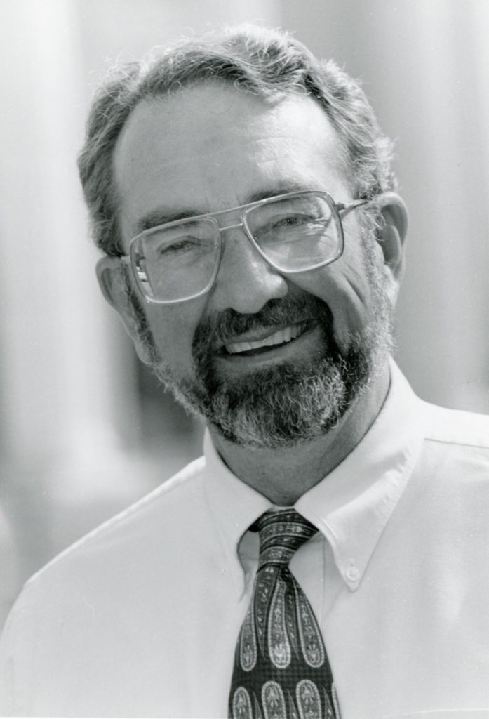 A black and white portrait picture of a man smiling brightly with a beard, and short hair and glasses wearing a white collared shirt and a patterned dark tie.