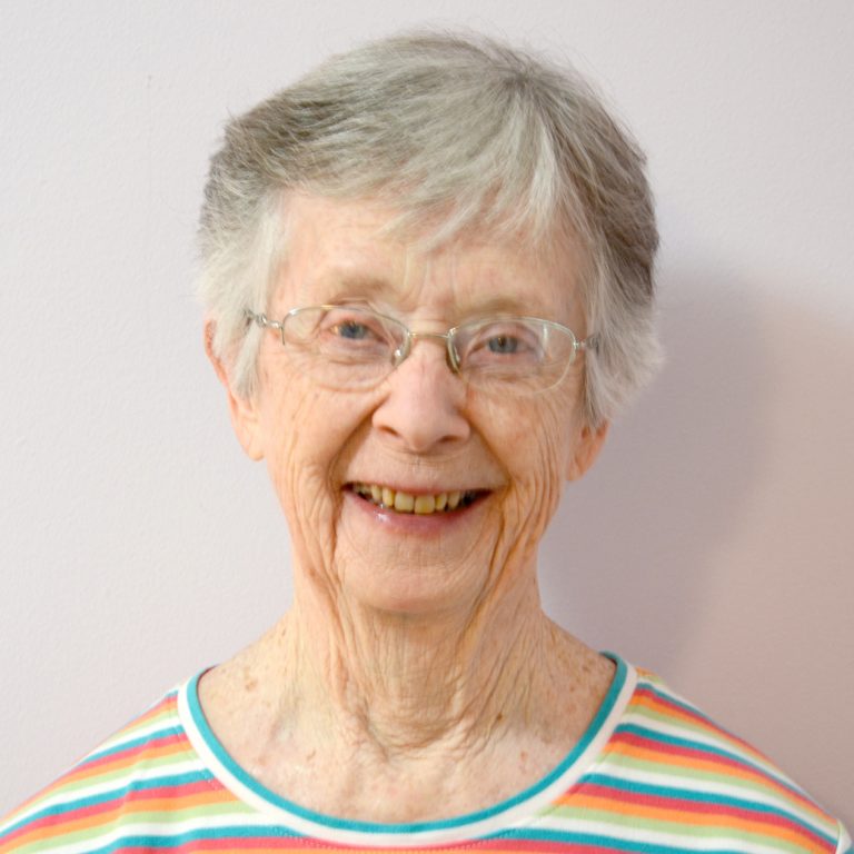 A woman with short, white and grey hair, wearing glasses and a striped colorful shirt smiles for a headshot picture gleefully in front of a plain white background.