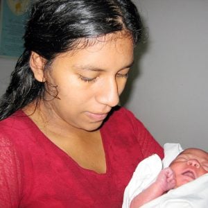 A young woman pictured candidly holding a newborn baby wrapped in a white blanket indoors.