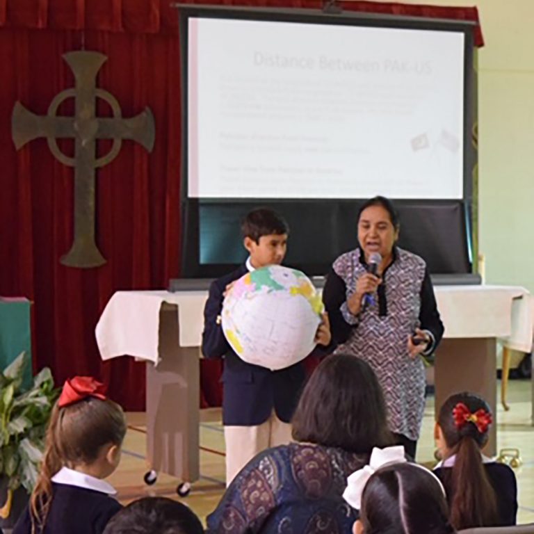 A young boy in a school uniform holding a paper lantern globe on stage with a teacher wearing a dress with a microphone talking to a crowd of young students with a projector and cross in the background indicating a religious school stage.