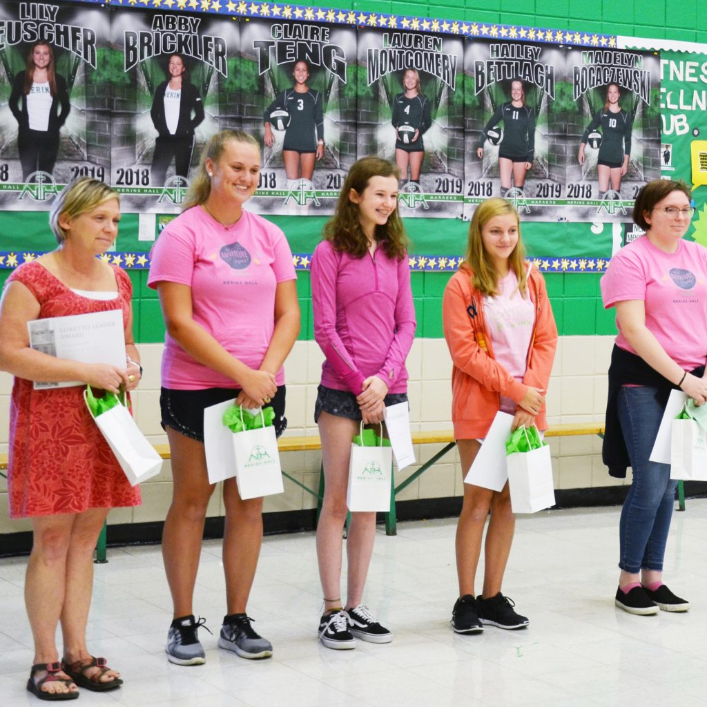 Five women standing in front of a high school gym with small white gift bags with bright green tissue paper in their hands.