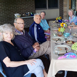 A group meal with all 8 smiling for a picture at a long outdoor table on a brick house patio.