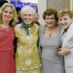 Four women smiling together for a group picture indoors while wearing dress clothes.