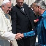 A man wearing a blue striped shirt shakes hands with the Pope outdoors.