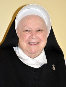 A nun smiling for a headshot picture with a plain yellow background.