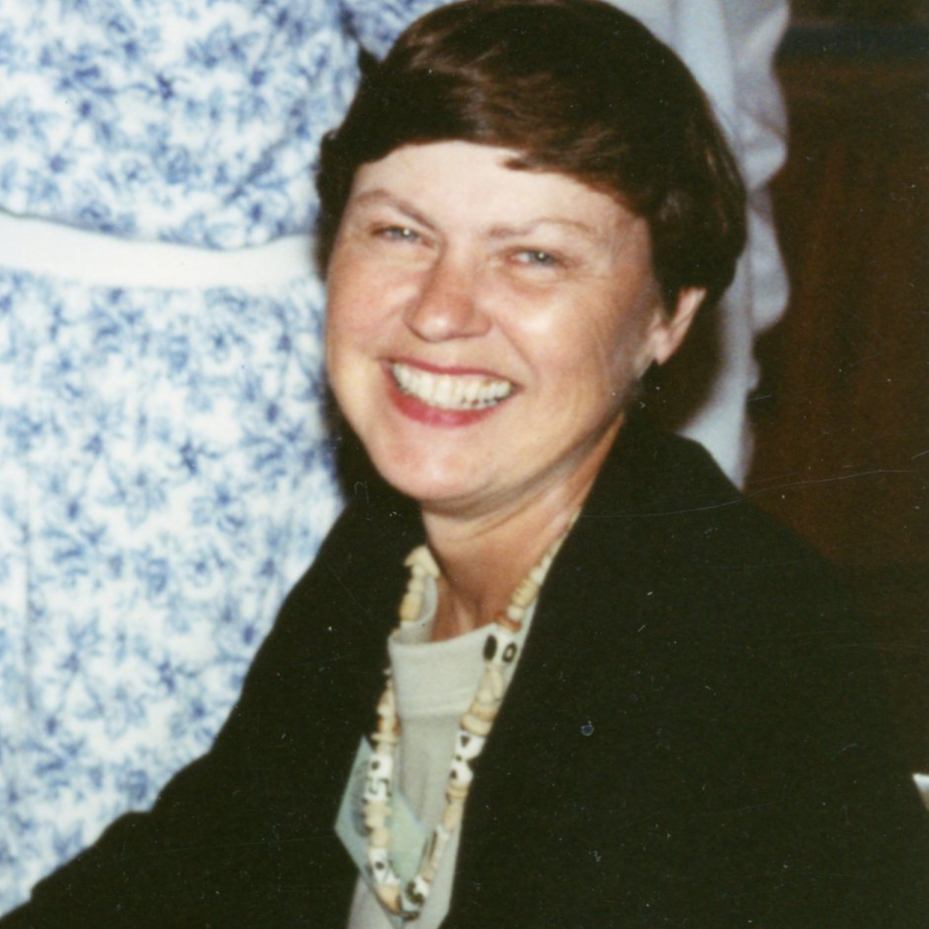 A woman with short brown hair smiling brightly wearing a beaded necklace, a beige blouse, and a black blazer indoors.