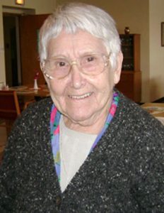 A woman with short white hair, big clear-framed glasses, wearing a white blouse, a patterned colorful scarf, and a grey cardigan sweater smiling for a headshot picture in a softly lit room.
