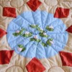 A close-up of a cream white quilt with a light blue circle with water lily patterns within and a border of eight red diamond patches surrounding the circle.