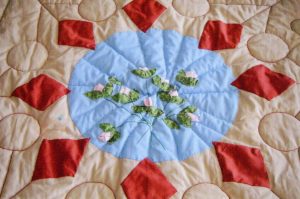 A close-up of a cream white quilt with a light blue circle with water lily patterns within and a border of eight red diamond patches surrounding the circle.