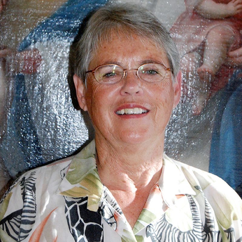 A woman with short grey hair smiling brightly for a headshot picture wearing oval wire-framed glasses and a jungle and animal patterned collared shirt.