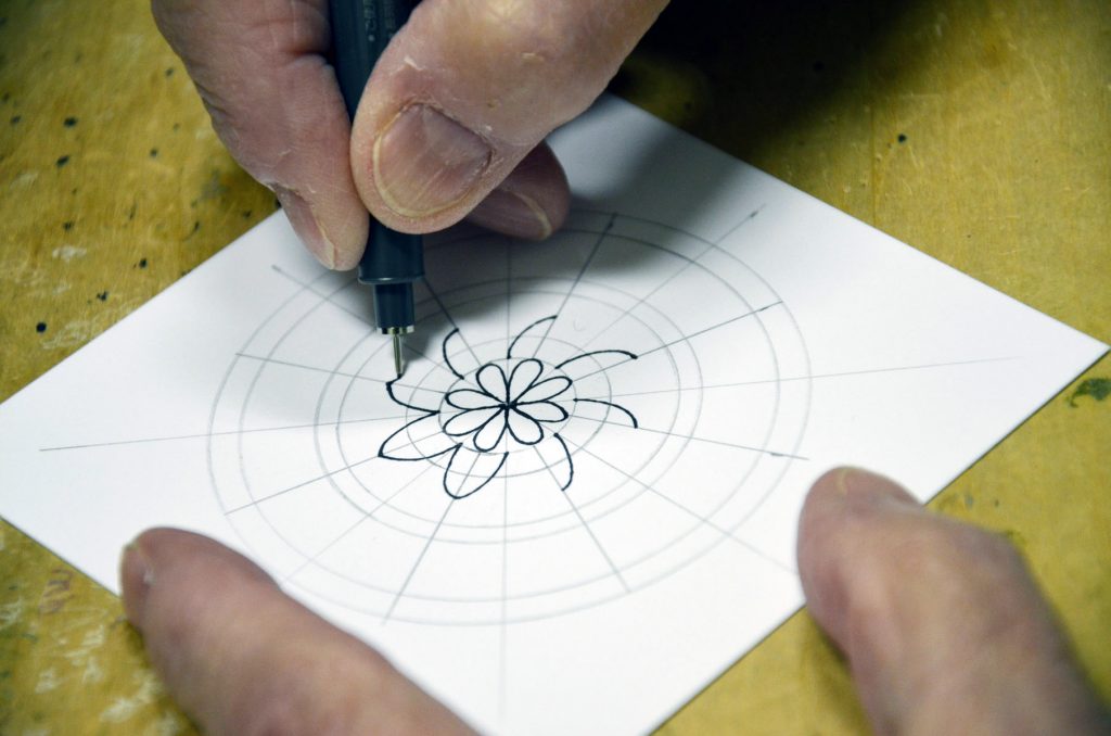 A mandala being created on a small square paper with a black pen on a wooden desk.