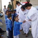 Three women wearing white outfits distributing water bottles to young students wearing blue school uniforms. They are outdoors in a shaded area on a sunny day. One woman wearing white is shaking a student's hand as he says thank you.