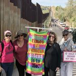 Protestors smiling for a picture at the U.S.-Mexico border. The protestors have bright signs and are wearing sunglasses.