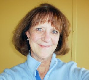 A woman with short brown hair and blue eyes smiling brightly for a selfie picture in front of a yellow wall.