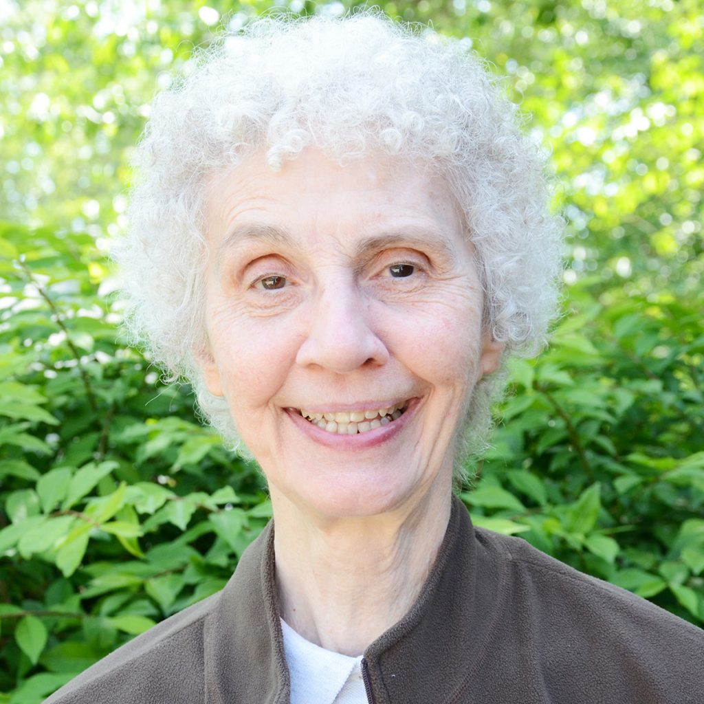 A woman with short, white hair, smiling brightly for a picture, wearing a brown jacket over a white t-shirt, with a leafy green background on a sunny day.