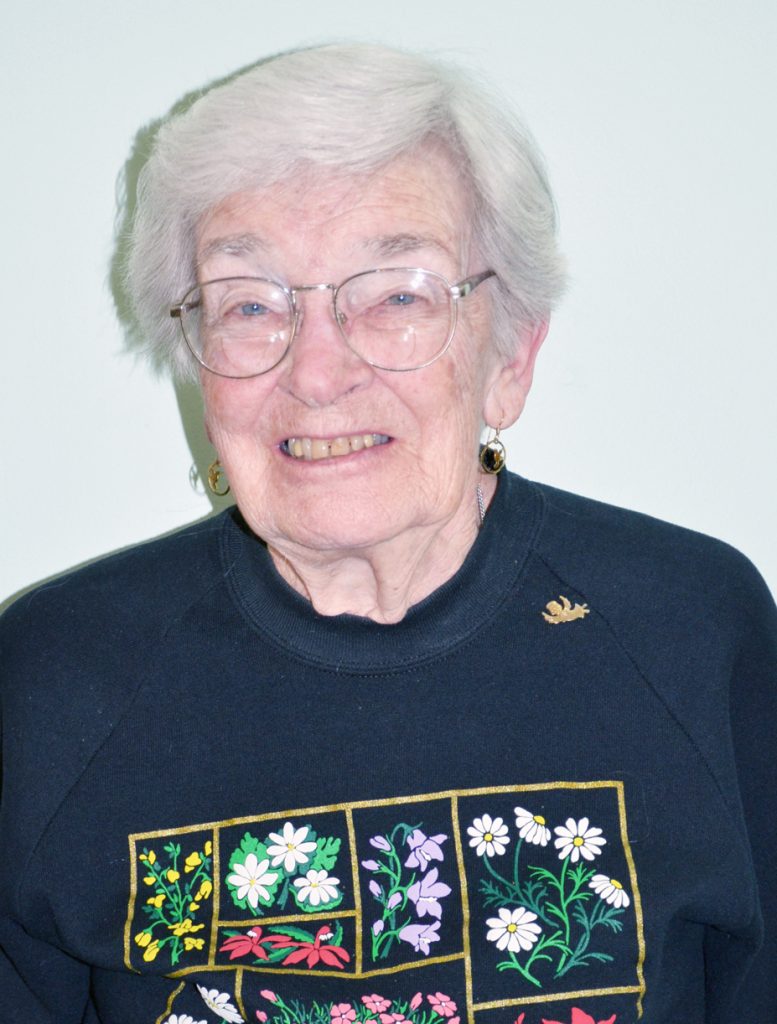 A headshot picture of a woman smiling with short, grey hair, wire glasses and a navy blue sweater with a floral grid print with a plain white background.