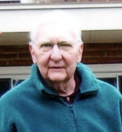 A man with short white hair, and glasses smiling for a picture wearing a greenish blue quarter zip.