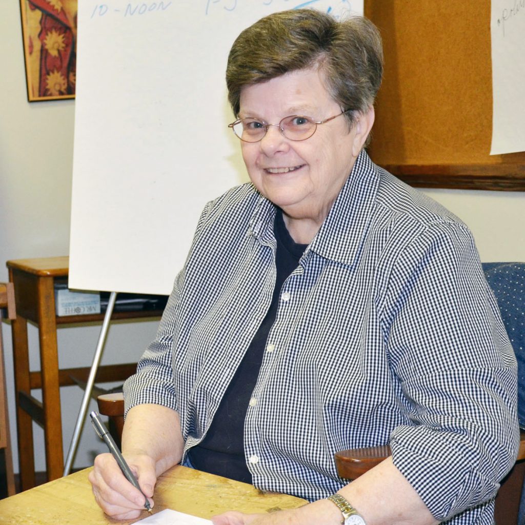 A woman with short brown hair and round wire-framed glasses smiling for a picture while drawing a mandala indoors.