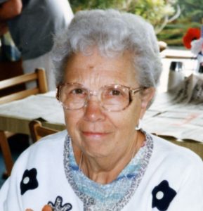 A woman with short, grey hair and glasses, smiling for a headshot picture outdoors, wearing a white patterned cardigan with black flowers.