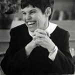 A black and white image of a woman with short dark hair, wearing a white collared shirt underneath a black sweater smiling joyfully in a candid picture with her hands clasped.